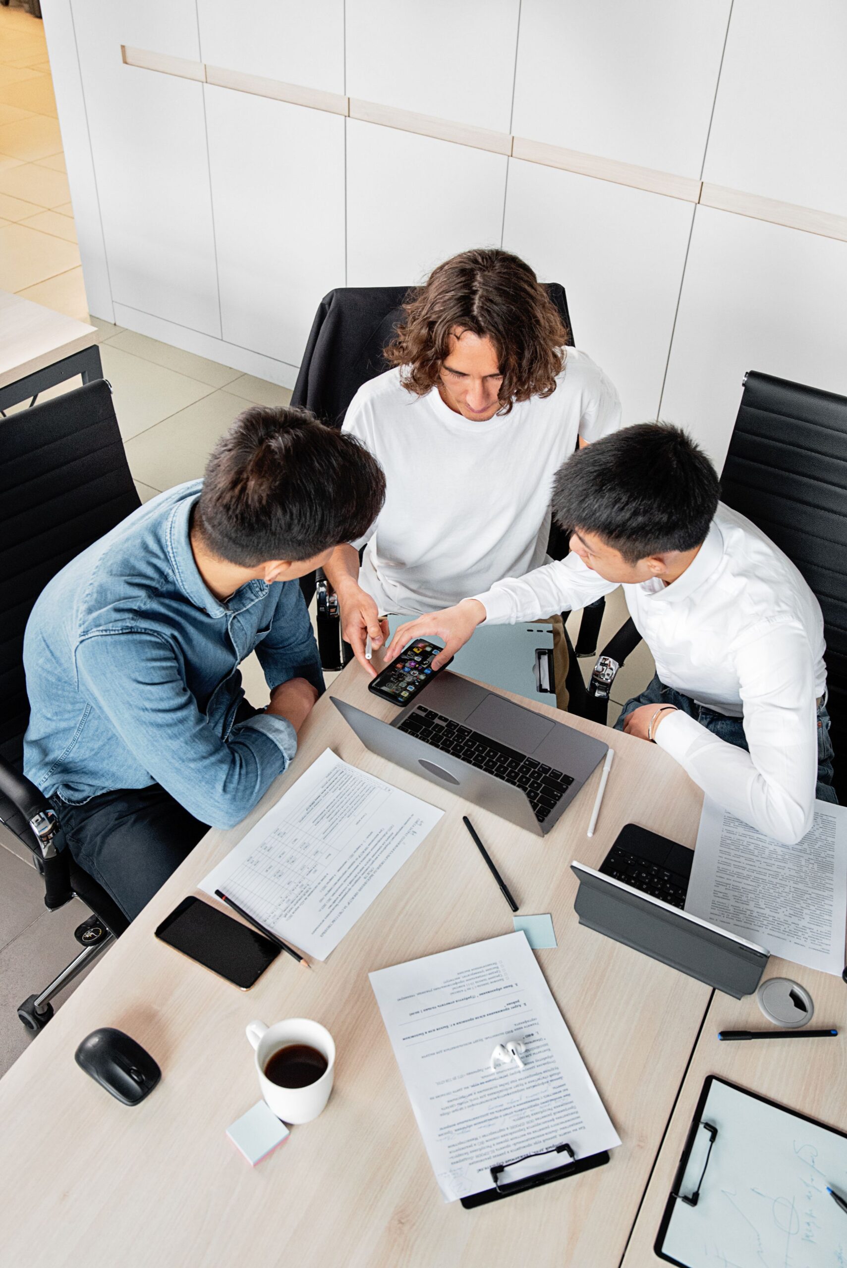 Three colleagues collaborating at a desk with laptops and documents, seen from above